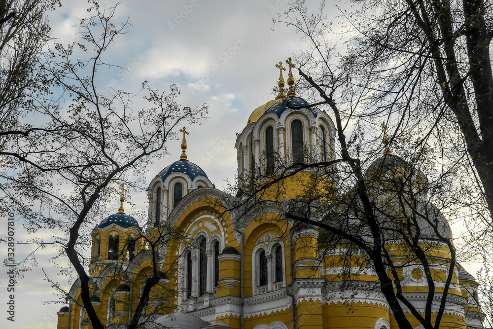 A view on St Volodymyr's Cathedral. The cathedral is painted yellow, with blue and golden-domed rooftop. View is disturbed by the tree branches. Overcast and cold day.