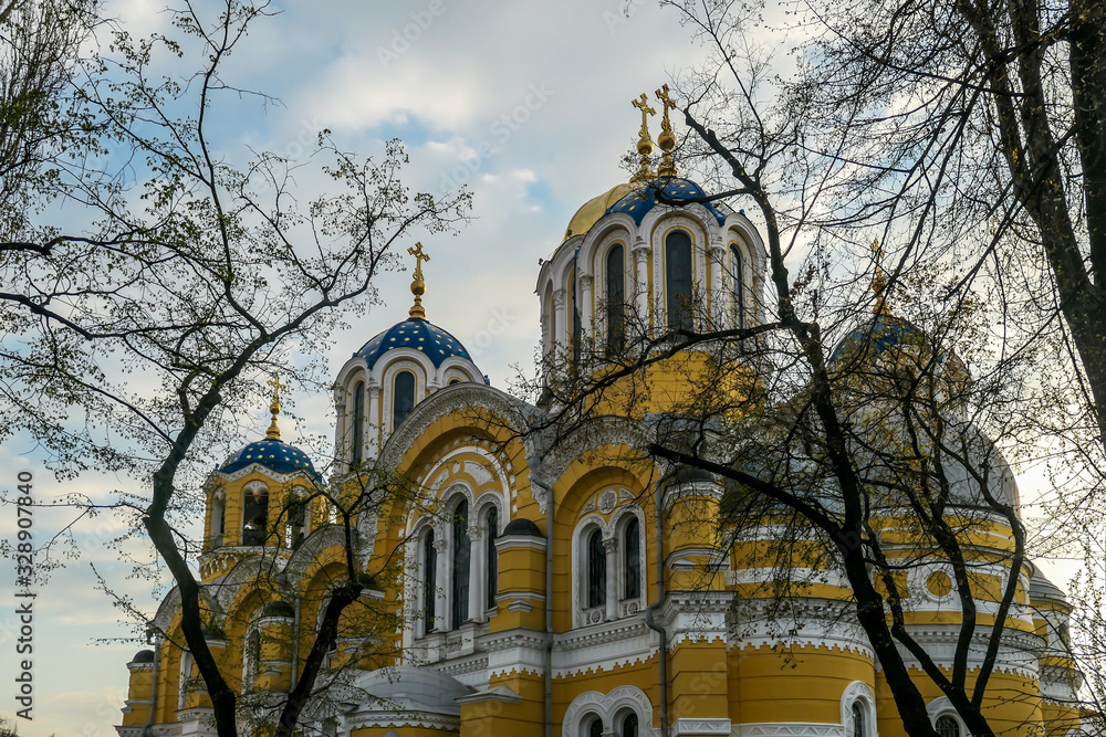 A view on St Volodymyr's Cathedral. The cathedral is painted yellow, with blue and golden-domed rooftop. View is disturbed by the tree branches. Overcast and cold day. A view on St Volodymyr's Cathedral. The cathedral is painted yellow, with blue and golden-domed rooftop. View is disturbed by the tree branches. Overcast and cold day.