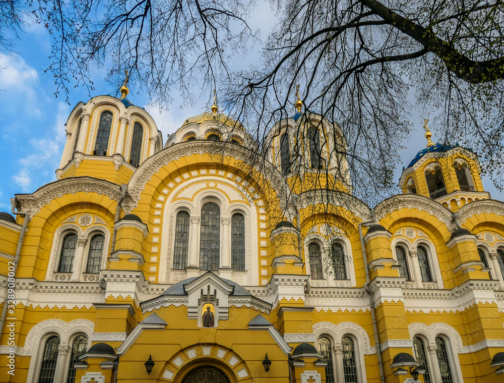 A view on St Volodymyr's Cathedral. The cathedral is painted yellow, with blue and golden-domed rooftop. View is disturbed by the tree branches. Overcast and cold day. A view on St Volodymyr's Cathedral. The cathedral is painted yellow, with blue and golden-domed rooftop. View is disturbed by the tree branches. Overcast and cold day.