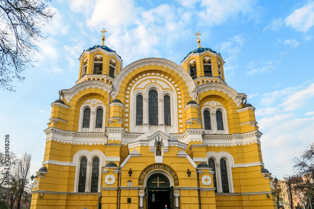 A view on the front facade of St Volodymyr's Cathedral. The cathedral is painted yellow, with blue and golden-domed rooftop. ON the side of the Cathedral there are trees. Overcast and cold day.