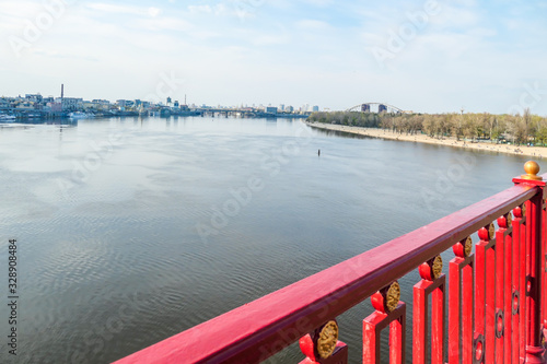 A view on Dnieper river from a pedestrian bridge. The balustrade is painted red. Bridge is leading to a small island in Kiev, which is a local beach. In the back there is a lot of tall buildings.