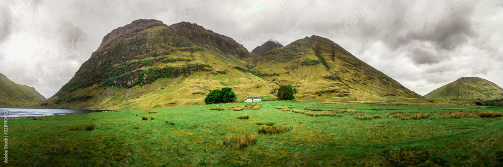 Achnambeithach cottage panorama in cloudy weather. Green scottish landscape. Famous house in northern Scotland. Beautiful ultra wide scotland highlands nature with cottage in the center. 