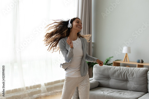 Overjoyed millennial girl wearing headphones have fun moving listening to music relax in living room, happy young woman in earphones dance enjoy leisure weekend at home, stress free concept