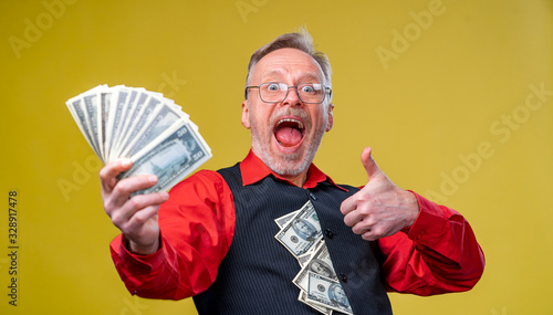 Old smiling grey-haired man in eyeglasses holding fan of dollars in front of the camera showing thumb up. Human emotions and facial expressions photo