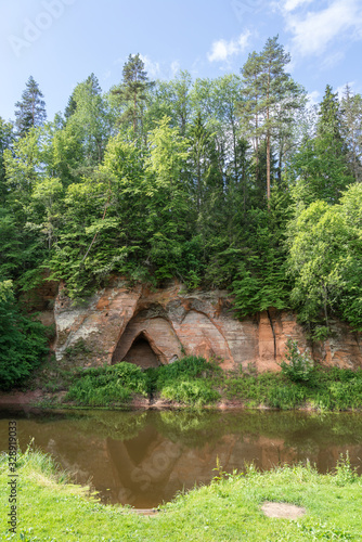 Sand stone cliffs and Salaca river. Red sandstones cave, water, trees and plants, green nature environment, climate concept..Angels cliff in Mazsalaca nature park, Latvia, Europe photo