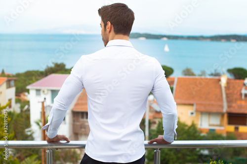 young man is wearing a white shirt and is looking at the sea view from the balcony.