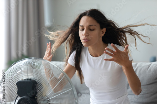 Millennial Caucasian girl sit on sofa in living room breathe fresh air from floor ventilator suffer from summer heat at home, young woman feel hot use fan at home struggle with lack of air condition