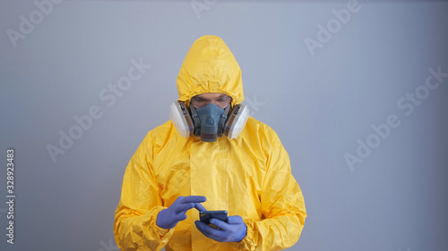 A man in yellow chemical protection suit and a respirator uses a phone gray background.