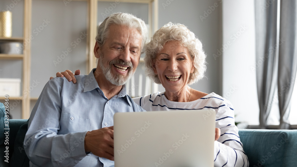 Overjoyed mature 60s husband and wife sit relax on sofa in living room have fun watch funny video on laptop, smiling elderly couple rest on couch browse internet use modern computer at home together Overjoyed mature 60s husband and wife sit relax on sofa in living room have fun watch funny video on laptop, smiling elderly couple rest on couch browse internet use modern computer at home together