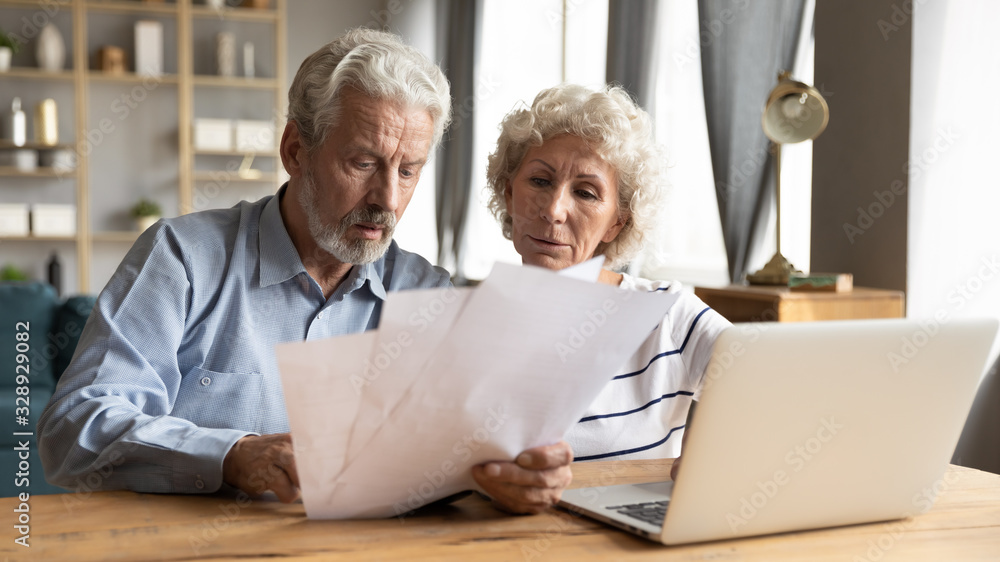 Focused elderly husband and wife sit at table in living room manage household document pay bills on laptop together, mature 60s couple spouses read bank contract use online system on computer Focused elderly husband and wife sit at table in living room manage household document pay bills on laptop together, mature 60s couple spouses read bank contract use online system on computer