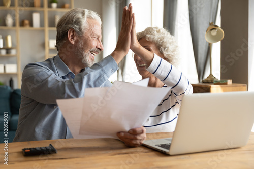 Happy old 60s couple spouses sit at desk at home give high five celebrate good news in letter document, smiling elderly husband and wife pay bills on internet on laptop online, easy banking system photo