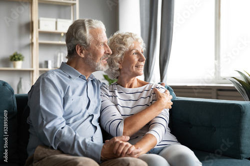 Optimistic elderly husband and wife sit on couch hugging look in distance dreaming or thinking of happy future together, smiling 60s old couple dreamers rest on sofa embrace cuddle visualize at home