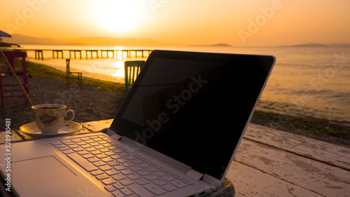 Freelance working with laptop on wooden table on beach at sunset. Woman sitting on chair and using computer near blue sea shore at sunny day. Digital nomadism towards yellow sky & horizon	