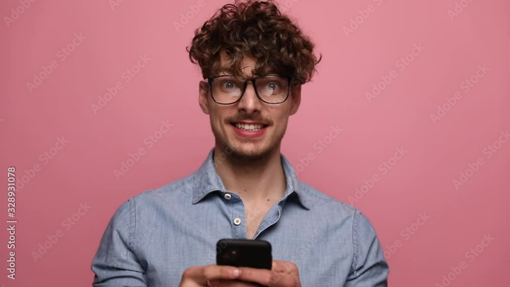 attractive casual man wearing glasses standing and holding his phone happy then raising one fist excited on pink studio background attractive casual man wearing glasses standing and holding his phone happy then raising one fist excited on pink studio background