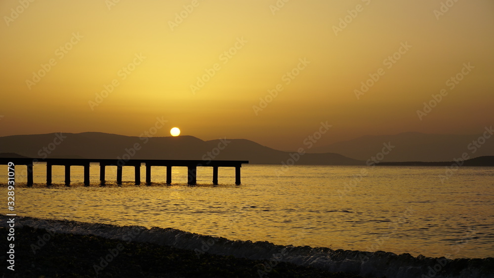 Dramatic sunset landscape at Urla, Izmir, Turkey. Beautiful blazing sunset over bright blue sea, pier, orange & purple sky above it with awesome golden rays of sun light reflection on calm waves.	
