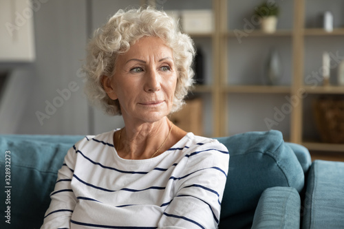Pensive old middle-aged 60s woman look in distance thinking pondering, thoughtful elderly lady sit relax on couch in living room daydreaming or visualizing, lost in thoughts missing mourning