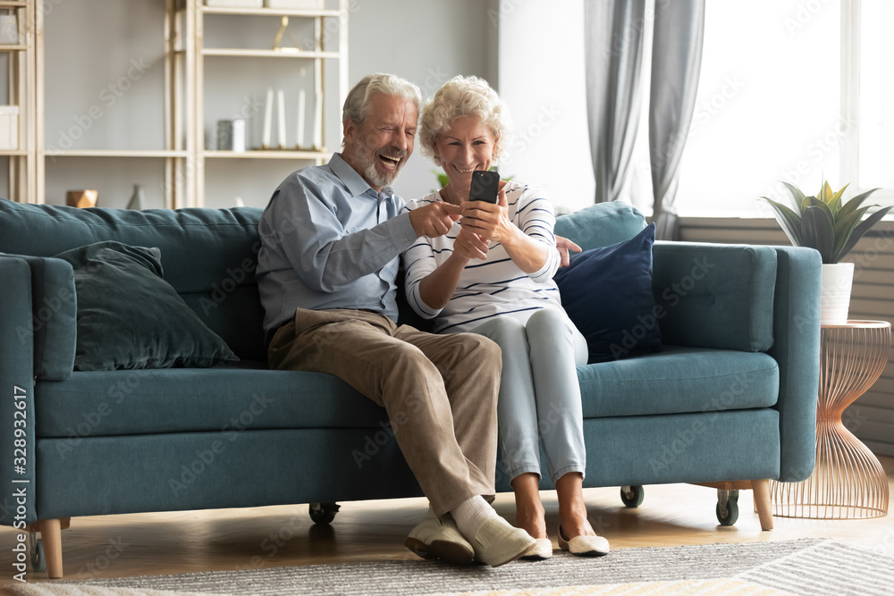 Happy middle-aged 50s husband and wife sit rest on couch in living room have fun making self-portrait picture on cell, excited elderly 60s couple laugh taking selfies using modern smartphone together