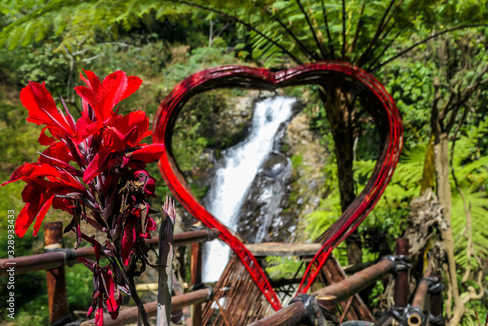 A red heart-shaped frame with Gitgit Twin Waterfall in the back, Bali Indonesia. There are red flowers. Touristic attraction for taking selfies with the waterfall. Lush green flora of the jungle.
