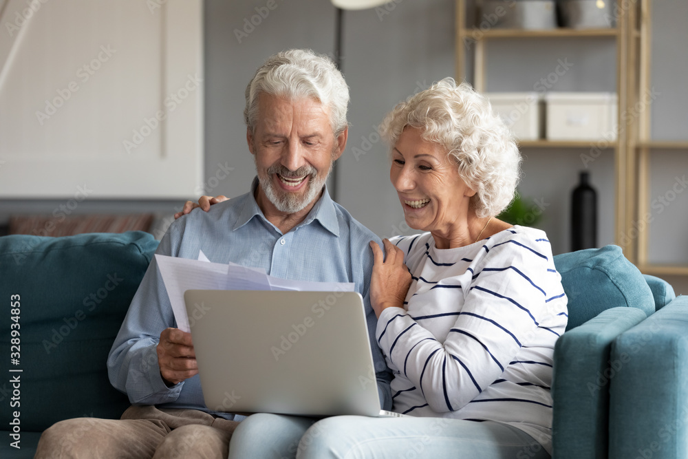 Happy elderly 60s couple sit rest on couch at home pay household expenses online on computer, smiling mature 50s husband and wife clients hold documents make payment on internet banking service