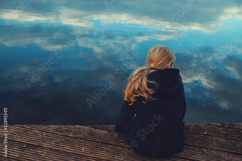 young girl with long blonde hair is sitting on a wooden stage while looking over the rippled water with reflecting cloudy sky in a thoughtful pose - coming of age, outdoor and nature care concept