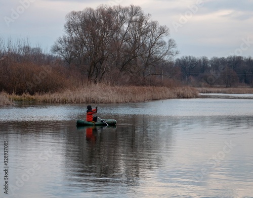 Spring scenery with a fisherman who fishes in an inflatable boat on the river, along the banks of which trees and dry reeds grow