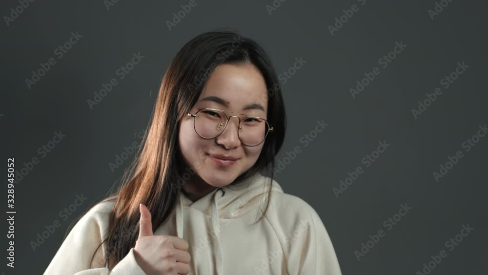 Young asian woman making thumbs up like sign over grey background. Winner. Success. Positive korean girl smiles to camera. Body language. Young asian woman making thumbs up like sign over grey background. Winner. Success. Positive korean girl smiles to camera. Body language.