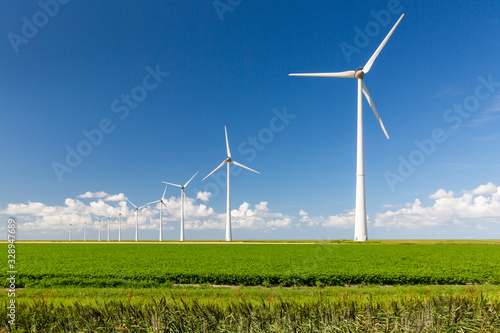 A long row of wind turbines emerging from green grass and against a summer blue sky with distant clouds