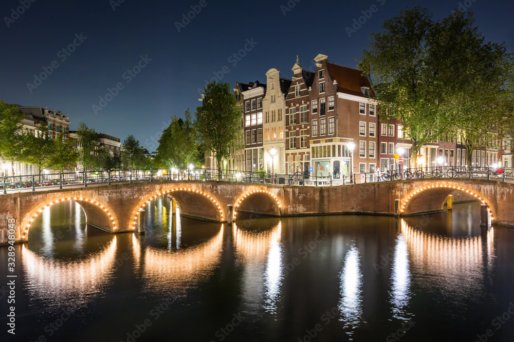 Wide angle cityscape of Amsterdam at night, with an historical bridge lit with nightlights reflecting on waters