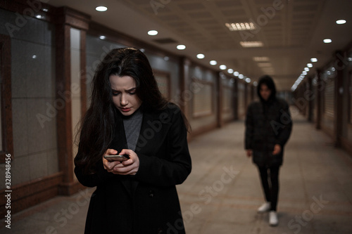 a young beautiful woman walks through an underground passage at night, followed by a man in dark clothing with a hood on his head. a woman tries to dial the number of the rescue service on the phone