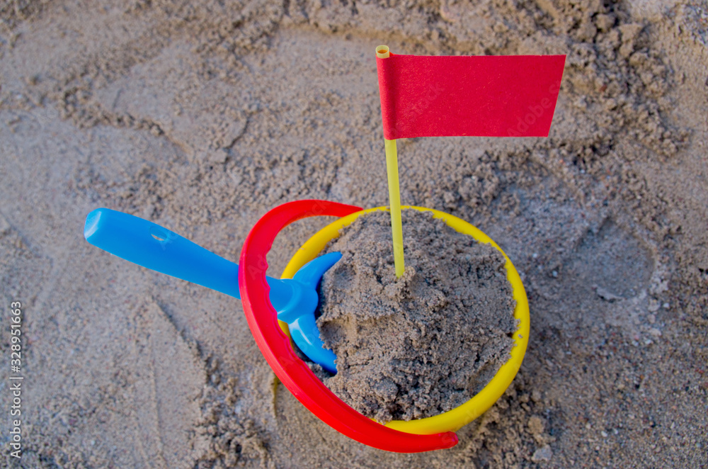 Red paper flag on yellow plastic flagpole rises above a pile of gray sand with a blue shovel stuck in the sand in a yellow bucket in sandbox. Concept: Reached goal, successfully completed worthy task Red paper flag on yellow plastic flagpole rises above a pile of gray sand with a blue shovel stuck in the sand in a yellow bucket in sandbox. Concept: Reached goal, successfully completed worthy task