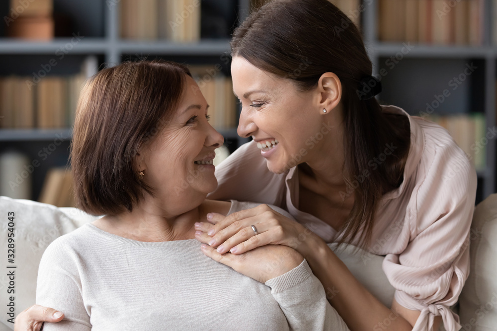 Laughing young woman embracing from back sitting on sofa smiling older mother. Affectionate grownup daughter sharing news, communicating talking to pleasant middle aged mature mommy at home. Laughing young woman embracing from back sitting on sofa smiling older mother. Affectionate grownup daughter sharing news, communicating talking to pleasant middle aged mature mommy at home.