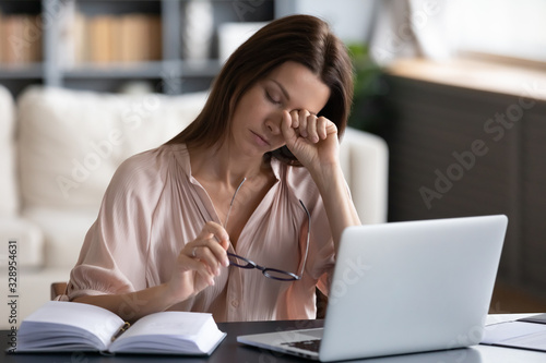Overwhelmed unhappy young woman taking off glasses, rubbing eyes, feeling exhausted, working remotely on computer at home. Tired stressed lady suffering from dry eyes syndrome, massaging nose bridge.