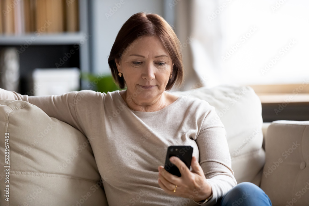 Worried middle aged woman sitting on couch, looking at mobile phone screen. Serious senior mature mother waiting for response from grownup children, chatting in social network online via smartphone. Worried middle aged woman sitting on couch, looking at mobile phone screen. Serious senior mature mother waiting for response from grownup children, chatting in social network online via smartphone.