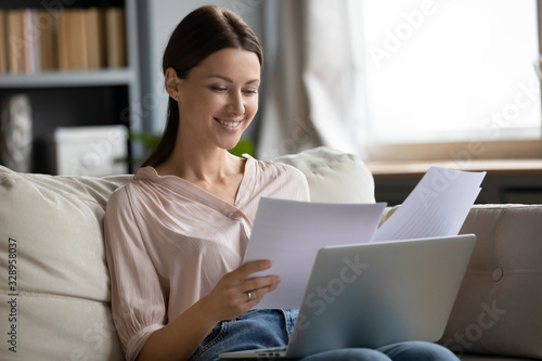 Smiling young woman sitting on sofa with computer, reading paper letters. Successful happy businesswoman working remotely at home, looking through documents, satisfied with financial reports.
