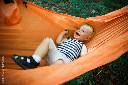 Happy, sincerely smiling little toddler boy lying in orange hammock in the forest emotional shot.