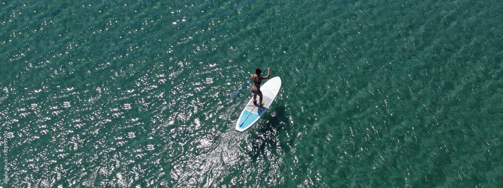 Aerial drone ultra wide photo of fit woman enjoying SUP or Stand Up Paddle board in shallow exotic bay with emerald sea