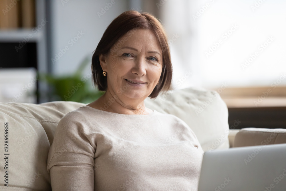 Head shot close up portrait smiling middle aged woman sitting on couch with laptop, looking at camera. Happy older senior grandmother using computer alone at home, retired people technology concept. Head shot close up portrait smiling middle aged woman sitting on couch with laptop, looking at camera. Happy older senior grandmother using computer alone at home, retired people technology concept.