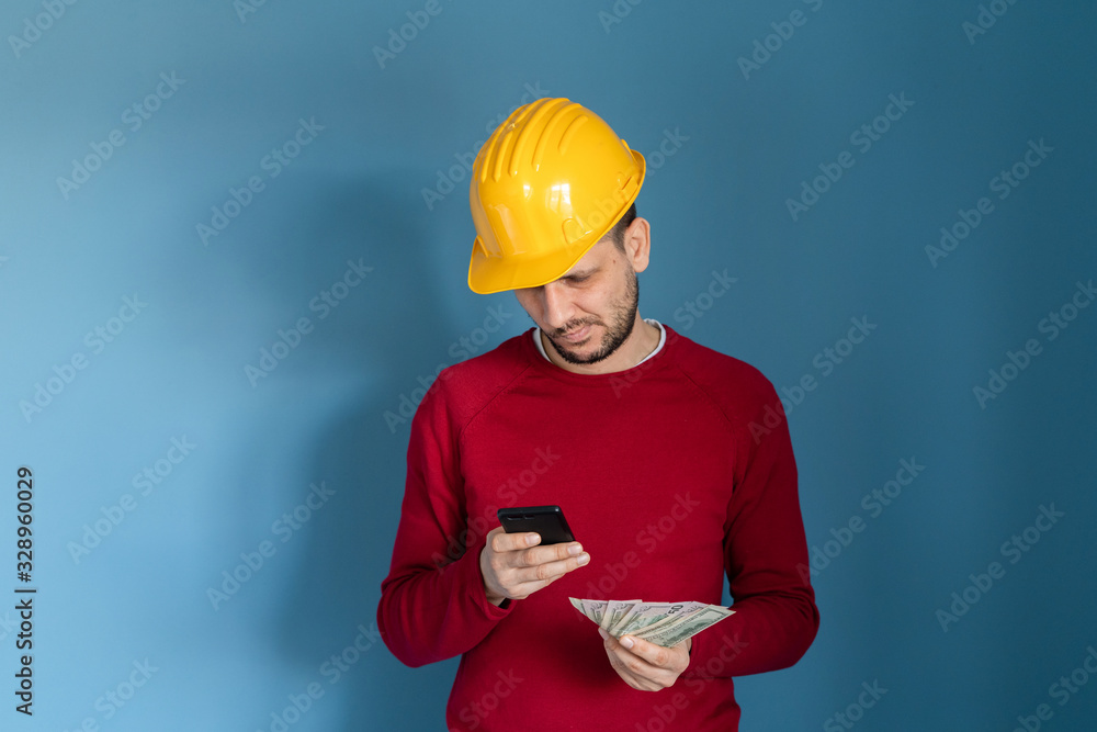 Portrait of young caucasian man wear red sweater in front of blue background with protective helmet construction engineer worker contractor holding money us dollar banknotes and mobile phone reading Portrait of young caucasian man wear red sweater in front of blue background with protective helmet construction engineer worker contractor holding money us dollar banknotes and mobile phone reading