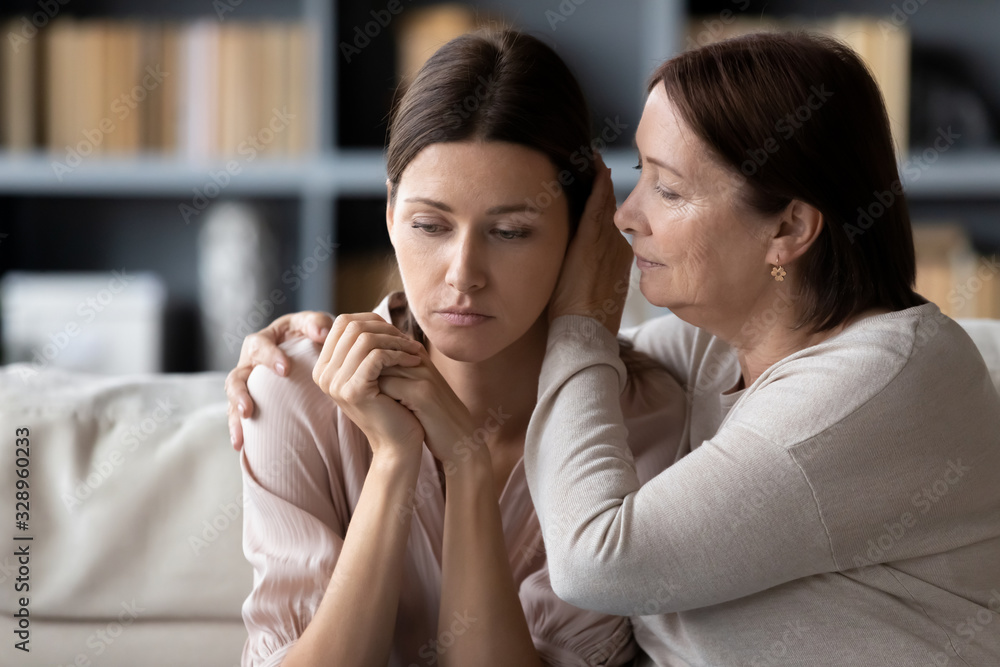 © fizkes - Compassionate middle aged mother comforting stressed upset daughter, sitting together on sofa at home. Kind senior mature mommy supporting soothing frustrated desperate unhappy grownup child.
