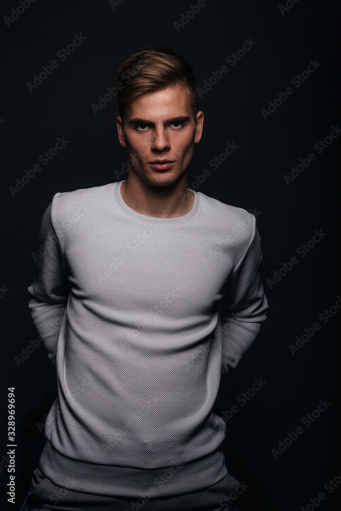 Informally ( casual ) dressed blonde young man with sharp jawline in his 20's posing in a studio in front of a black background while wearing a white sweater.