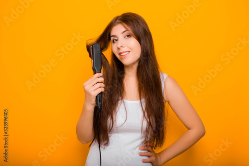 Attractive woman in white clothes straightens hair with an iron, standing on a yellow background.