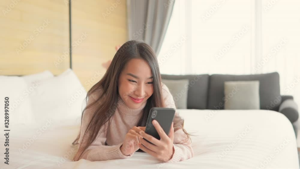 Attractive young Asian woman laying on large comfortable bed with smartphone in hand. Lady smiling while texting and relaxing in luxurious resort suite. Attractive young Asian woman laying on large comfortable bed with smartphone in hand. Lady smiling while texting and relaxing in luxurious resort suite.