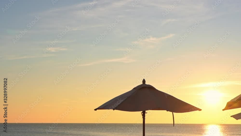 Stunning sunset sky and reflection on flat calm ocean surface behind open beach umbrellas. Tropical island luxury resort with colors of setting sun and soft clouds. PAN RIGHT. Stunning sunset sky and reflection on flat calm ocean surface behind open beach umbrellas. Tropical island luxury resort with colors of setting sun and soft clouds. PAN RIGHT.