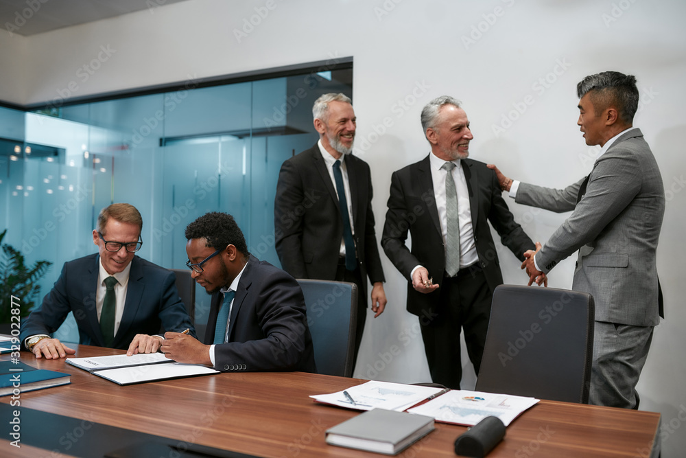 My congratulations. Two happy mature businessmen in classic wear shaking hands while having a multi-ethnic meeting in the modern office My congratulations. Two happy mature businessmen in classic wear shaking hands while having a multi-ethnic meeting in the modern office