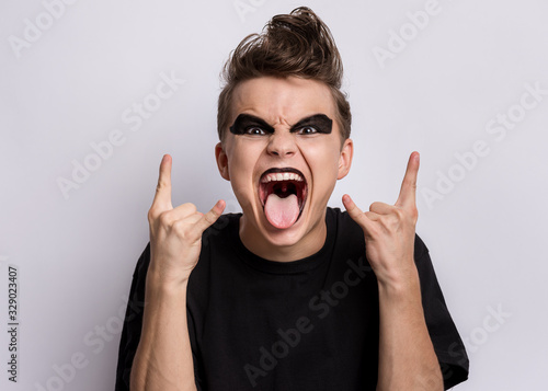 Crazy teen boy with spooking make-up making Rock Gesture on gray background. Teenager in style of punk goth dressed in black shows tongue and doing heavy metal rock sign. Problems of transitional age