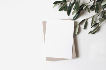 Summer wedding stationery mock-up scene. Blank vertical greeting card, craft paper envelope and olive branches isolated on white table background. Feminine Mediterranean flat lay, top view.