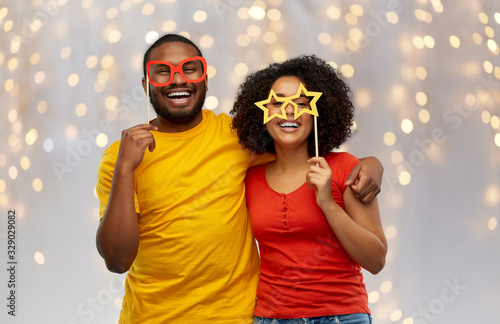 holidays, photo booth and people concept - happy african american couple with party props hugging over festive lights background photo