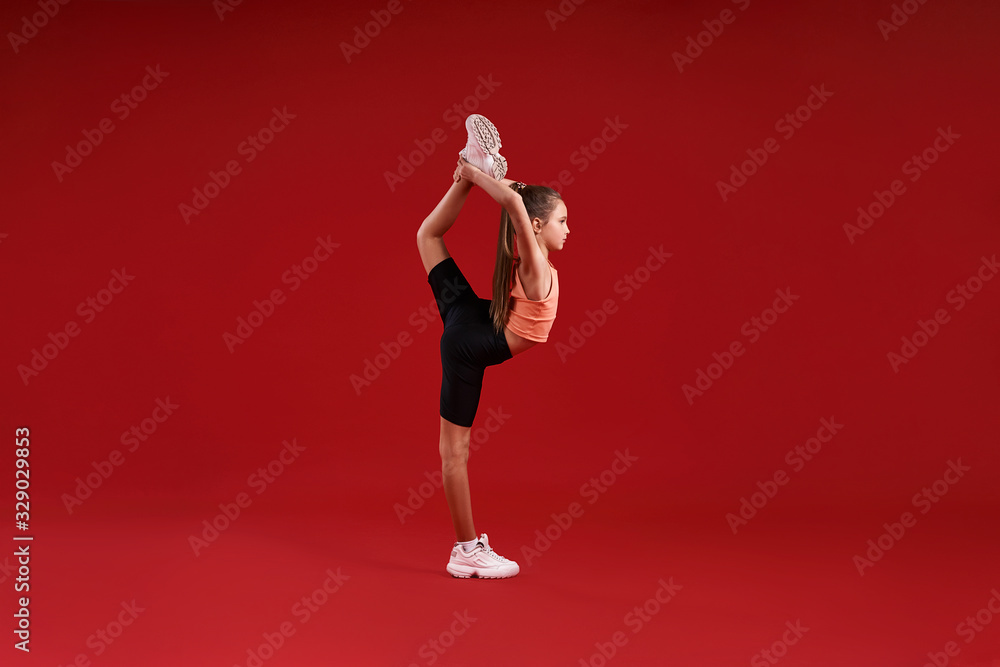 Excellence. A cute kid, girl is engaged in sport, gymnast is looking aside while stretching her legs. Isolated on red background. Fitness, training, active lifestyle concept. Horizontal shot