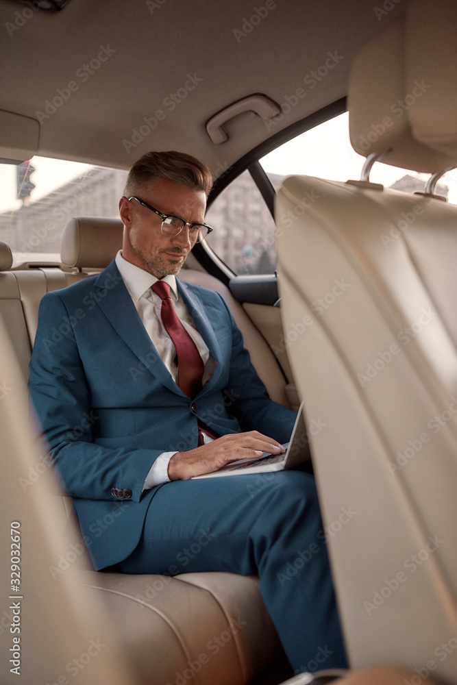 Never stop working. Serious mature businessman in full suit working on his laptop while sitting in the car