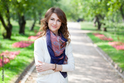 beautiful young brunette smiling woman in white jumper and with blue scarf in spring green park. alley with trees and flowers are in background. girl's arms crossed on her chest in a closed position